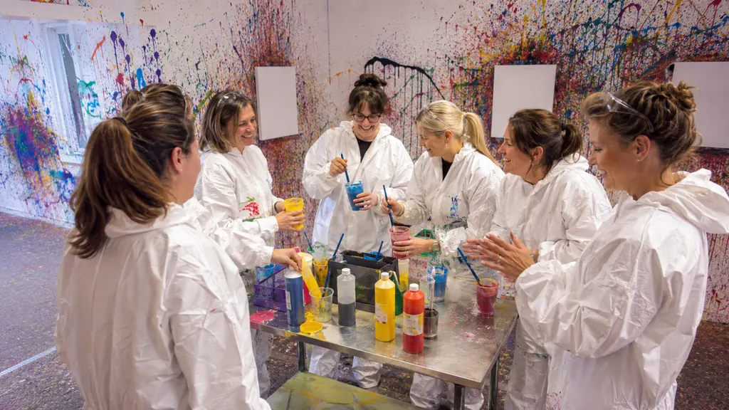 Kolleginnen in weißen Overalls mischen Farben in Bechern, an der Wand hinter ihnen bunte Farbspritzer