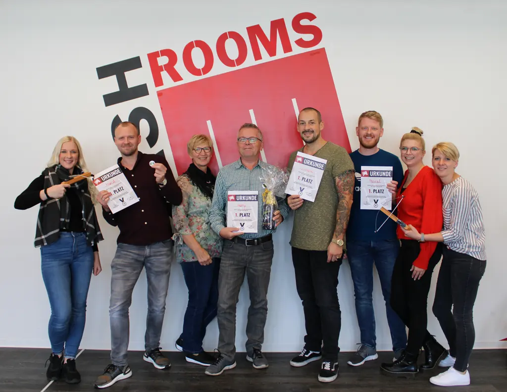 Eine gemischte Gruppe hält lachend ihre Urkunden vor einer weißen Wand