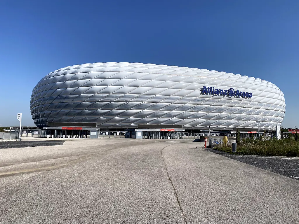Die weiße Allianz Arena mit ihrer rautenförmigen Kissenfassade steht unter tiefblauem, wolkenlosem Himmel, davor liegt ein breiter, leerer Fußweg.