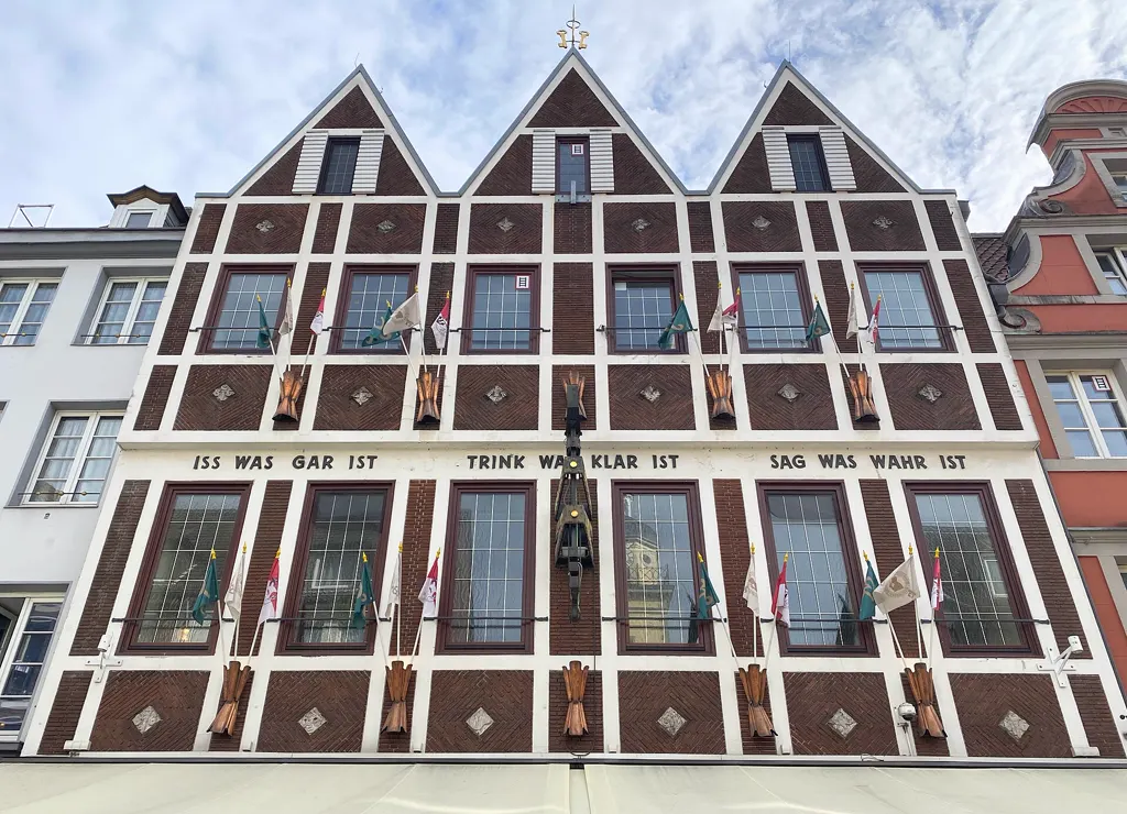 Fassade der Hausbrauerei Zum Schlüssel in der Düsseldorfer Altstadt mit drei Giebeln, Fahnen und dem Spruch „Iss was gar ist, trink was klar ist, sag was wahr ist“.