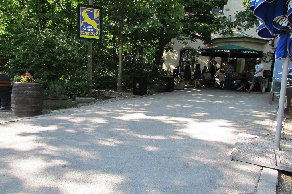 Der Hauptweg im Biergarten des Augustiner-Kellers unter Kastanienbäumen, hinten das Kellergebäude mit grünem Schirm und einigen Gästen, links ein Holzfass.