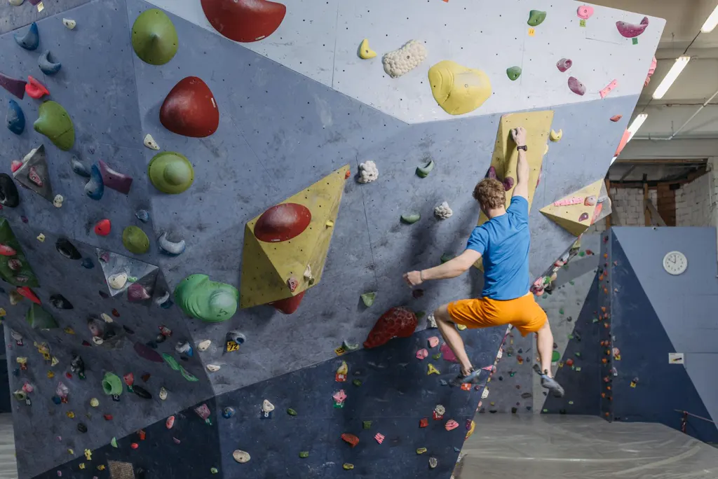 Ein Boulderer in blauem Shirt und gelber Hose greift, von hinten gesehen, an einer überhängenden Wand mit großen bunten Griffen.