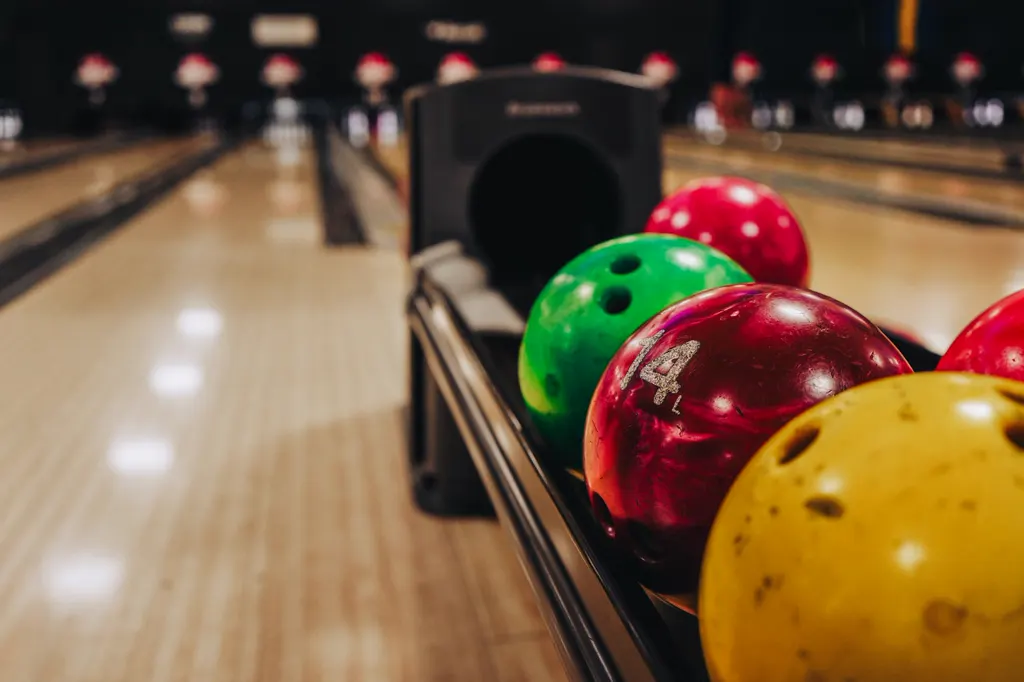 Bunte Bowlingkugeln im Ballrücklauf, dahinter helle Holzbahnen mit Pins, warmes Licht