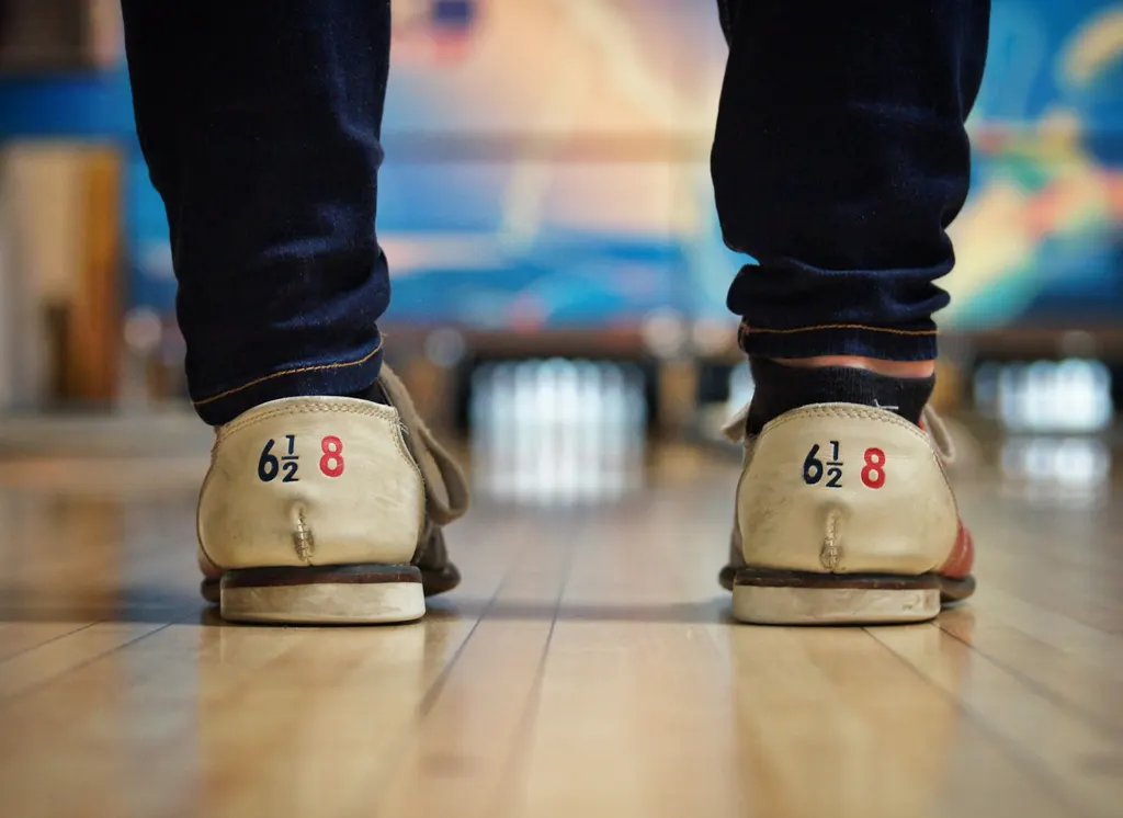Bowling-Leihschuhe mit Größenaufdruck von hinten auf einer glänzenden Holzbahn, im Hintergrund unscharf die Pins und eine bunte Wanddeko.