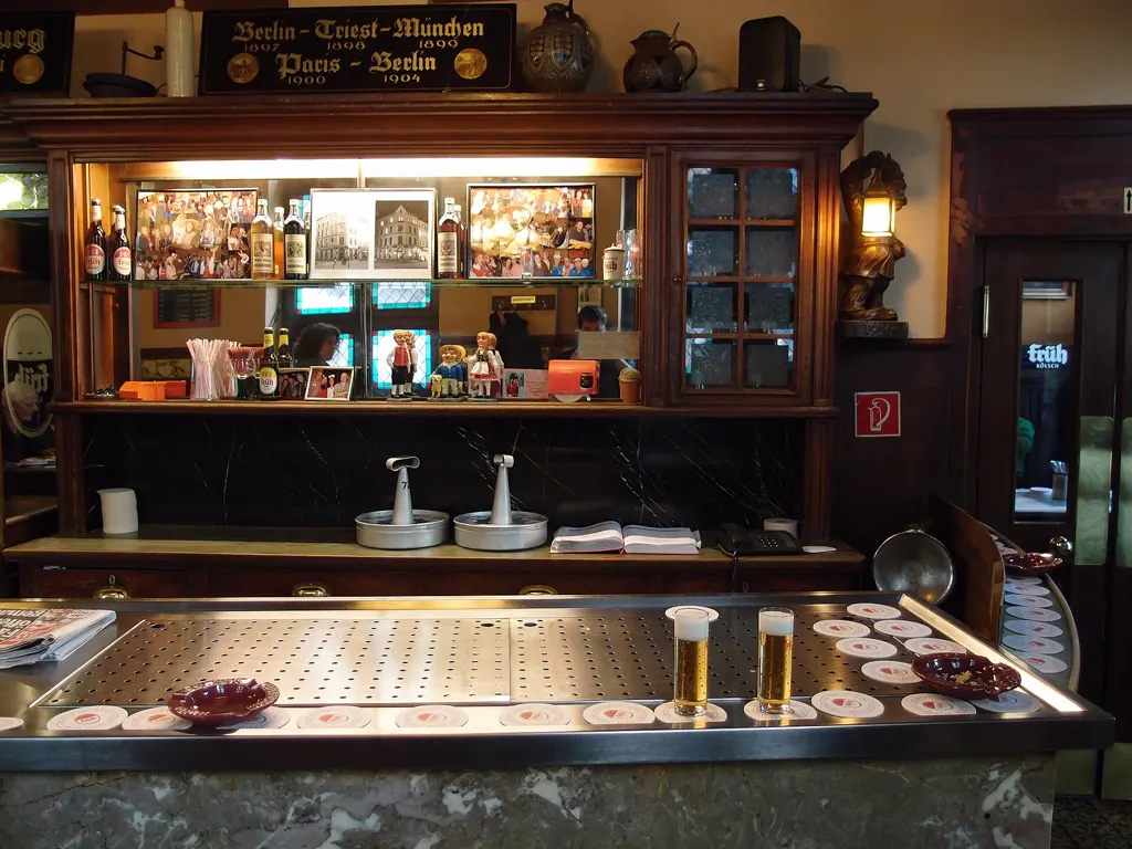 Holztheke im Kölner Brauhaus Früh em Veedel mit zwei frisch gezapften Kölsch-Stangen auf dem Edelstahl-Tresen, dahinter ein Buffetschrank mit Krügen und Flaschen.