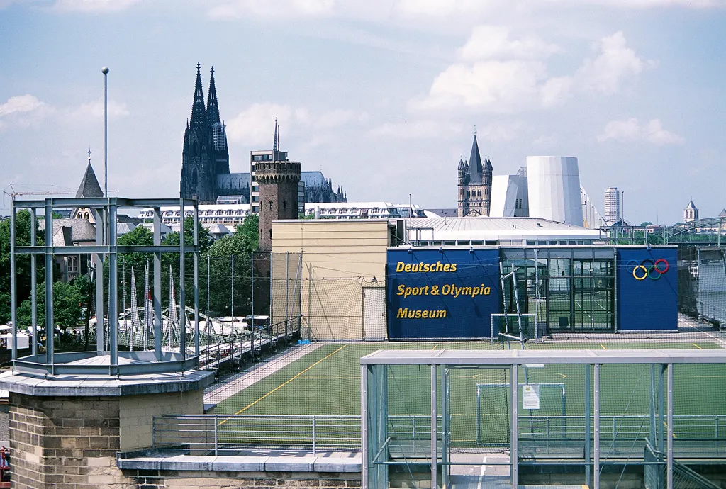Kunstrasen-Sportplatz auf dem Museumsdach mit dem Schriftzug „Deutsches Sport & Olympia Museum“ und den olympischen Ringen, dahinter Kölner Dom und Groß St. Martin unter blauem Himmel.