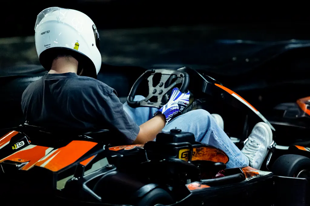 Kartfahrer mit weißem Helm, von hinten fotografiert, lenkt ein orange-schwarzes Leihkart auf einer dunklen Indoor-Bahn.