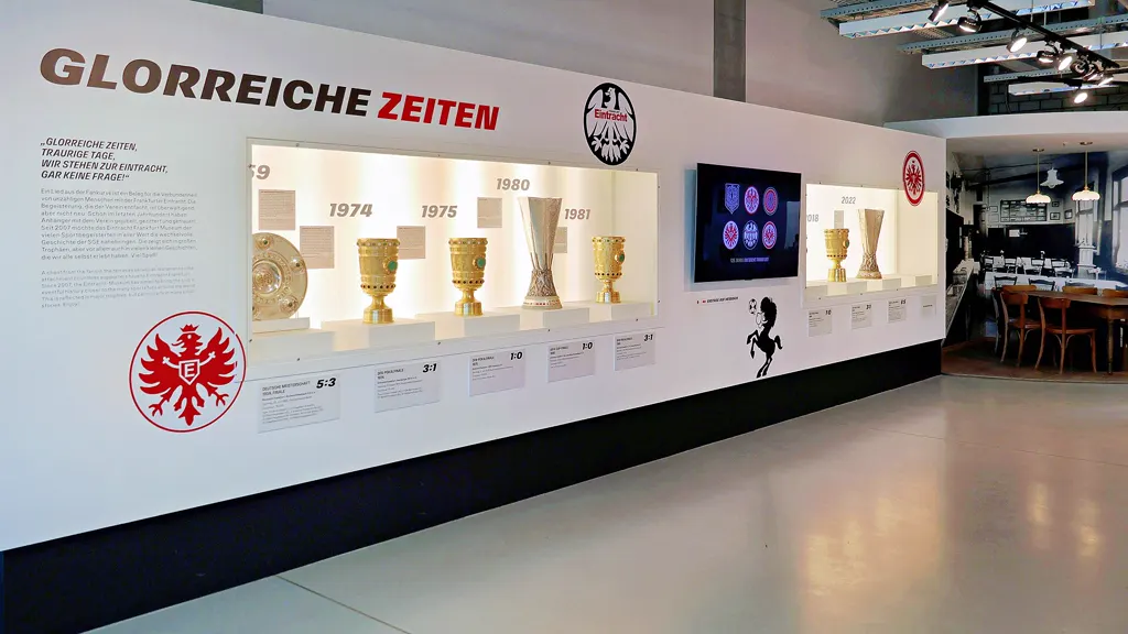 Ausstellungswand „Glorreiche Zeiten“ im Eintracht Frankfurt Museum mit Pokalvitrine, Eintracht-Adler und Bildschirm.