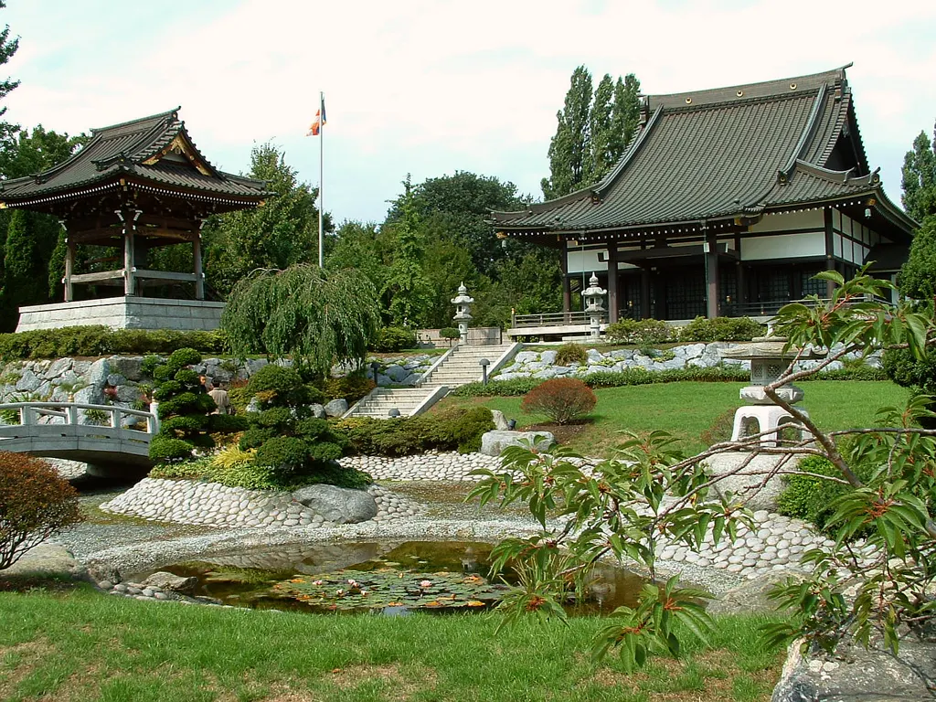Farbiger japanischer Garten des EKŌ-Hauses mit Teich, Steinen und Steinlaterne, dahinter Glockenturm und Tempel unter leicht bewölktem Himmel.
