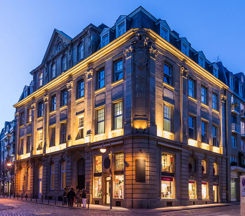Das Farina-Haus an der Ecke Obenmarspforten in der blauen Stunde: warm angestrahlte Sandsteinfassade, an der Hausecke die Inschrift mit dem Gründungsjahr 1709, erleuchtete Schaufenster, vor dem Eingang eine kleine Gruppe Menschen.