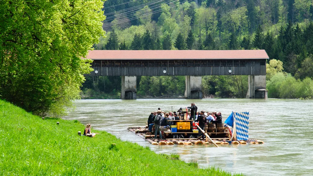 Ein Holzfloß mit einer Gruppe, Bierfässern und weiß-blauer Fahne treibt auf der Isar unter einer überdachten Holzbrücke hindurch, links eine sonnige grüne Uferwiese.