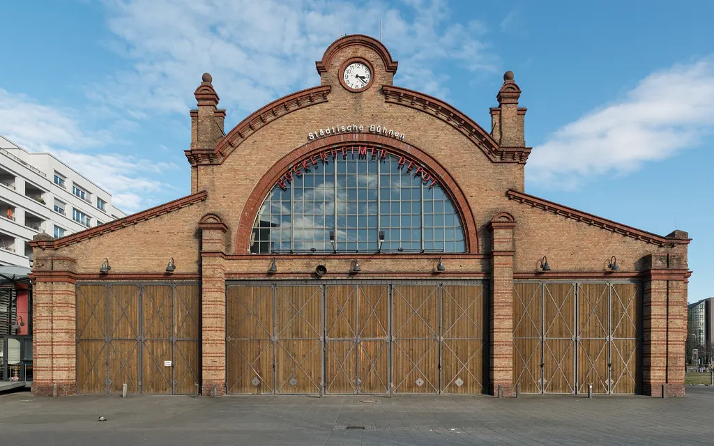Backsteinfassade des Bockenheimer Depots mit Uhr, großem Rundbogenfenster und Holztoren vor blauem Himmel.
