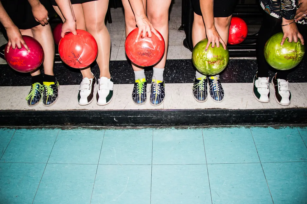 Fünf Personen stehen in Leih-Bowlingschuhen nebeneinander an der Bahn und halten bunte Kugeln in Pink, Rot und Grün. Im Bild sind nur Beine und Hände, darunter türkisfarbener Fliesenboden.