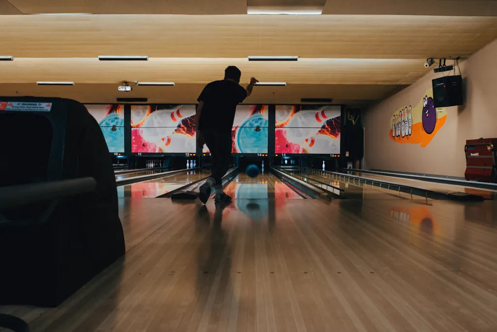 Helle Bowlinghalle mit langen Holzbahnen; ein Spieler wirft, von hinten als Silhouette zu sehen, die Kugel rollt auf die Pins zu, darüber bunte Bahnabdeckungen.