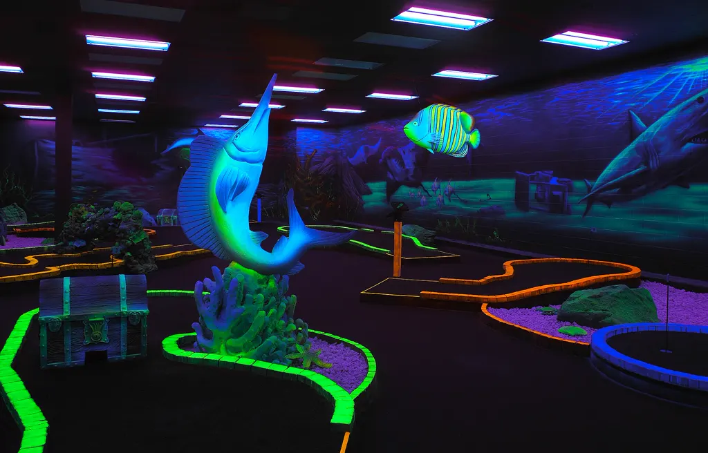 Leere Schwarzlicht-Minigolfhalle im Unterwasser-Stil: leuchtend grüne und orange Bahnbegrenzungen, Schwertfisch-Figur auf Korallen, Schatztruhe, schwebender Kaiserfisch und Wandbild mit Hai unter UV-Röhren.
