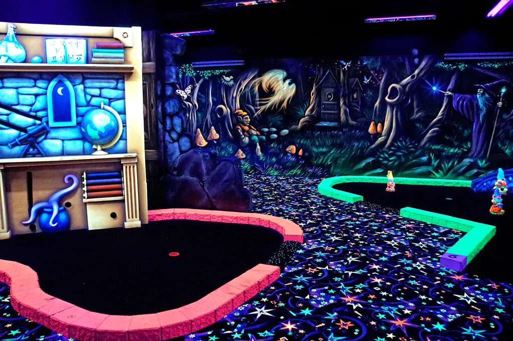 Schwarzlicht-Minigolfbahn im Zauberer-Stil: Bahn mit pink leuchtender Begrenzung und Loch, Teppich mit Leuchtsternen, grün leuchtende Bahn im Hintergrund, links ein Regal mit Globus und Büchern, an der Wand ein Waldbild mit Zauberer.