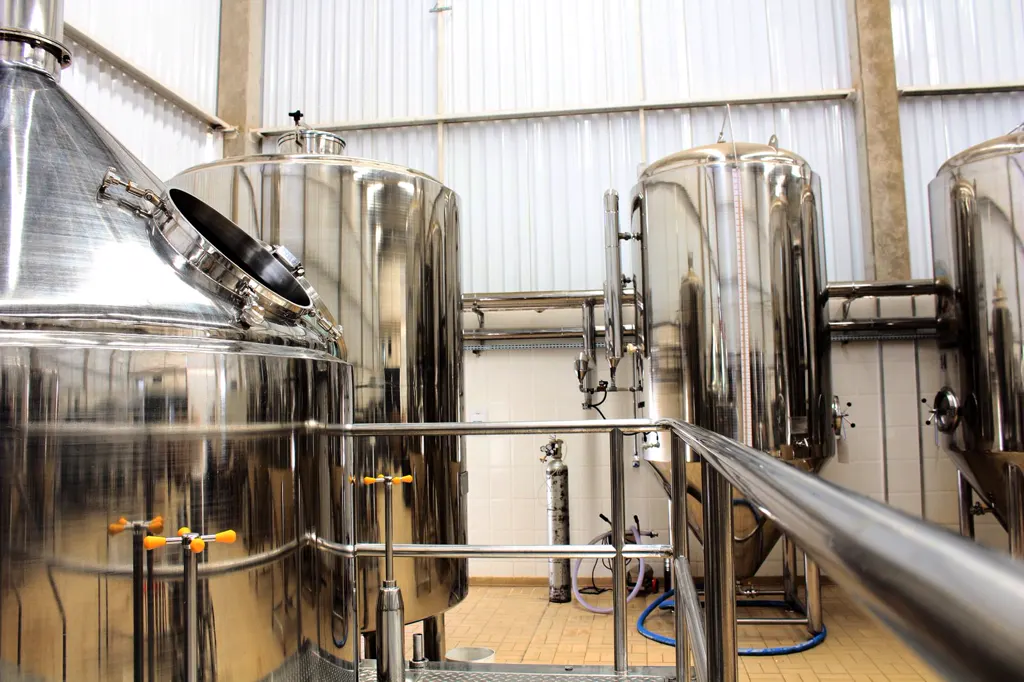 Glänzende Sudkessel und Gärtanks aus Edelstahl stehen in einem hellen, kleinen Brauhaus, vorne ein Edelstahlgeländer.