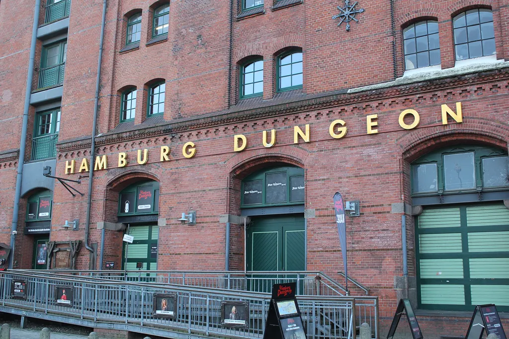 Backsteinfassade des Speichers am Kehrwieder mit goldenem Schriftzug „Hamburg Dungeon“, grünen Toren, Rampe und Geländer bei Tageslicht.