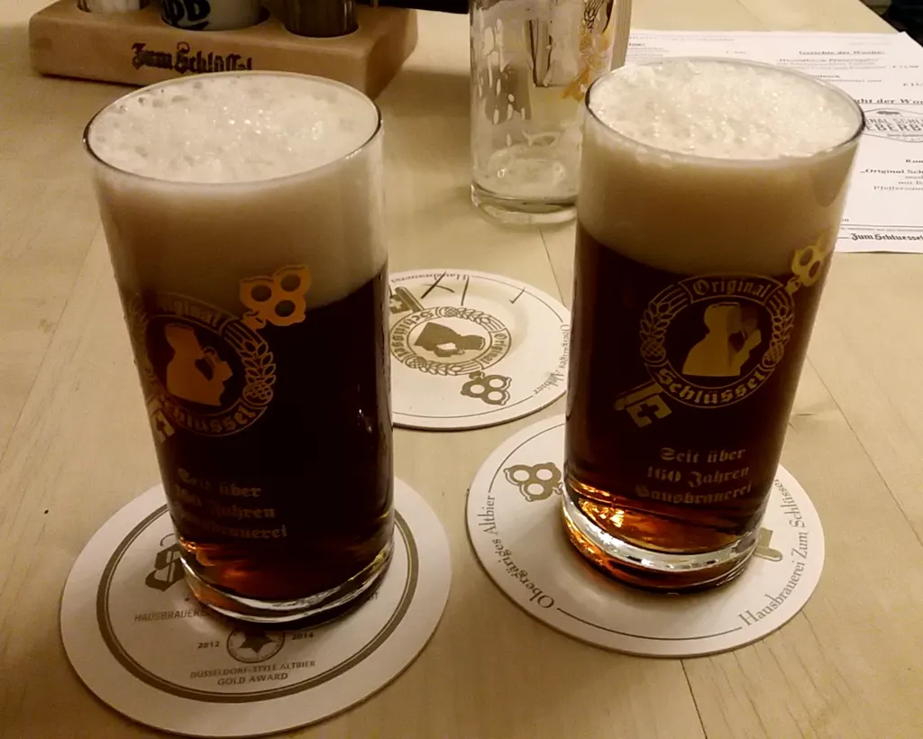 Zwei Gläser Original Schlüssel Altbier auf Bierdeckeln am Brauhaustisch, mit dem Hauslogo auf Gläsern und Deckeln