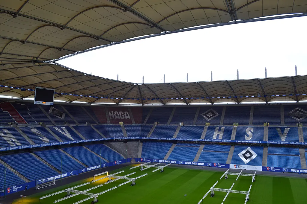 Blick von der Tribüne ins leere Volksparkstadion: blaue Sitzreihen mit dem HSV-Schriftzug und der Raute, darüber das Membrandach, auf dem Rasen stehen Belichtungsanlagen.