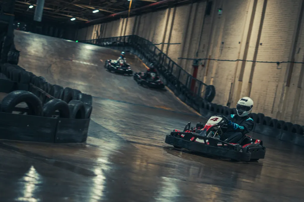 Drei Leihkarts mit behelmten Fahrern fahren über eine überhöhte Indoor-Kartbahn in einer Backsteinhalle.