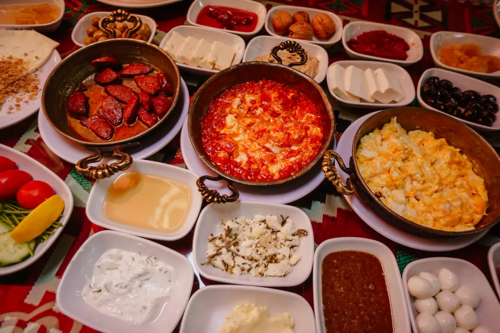 Türkisches Frühstück auf einem rot gemusterten Tischtuch: Sucuk, Menemen und Rührei in Kupferpfännchen, rundherum viele Schälchen mit Käse, Oliven, Walnüssen, Wachteleiern, Joghurt und Aufstrichen.