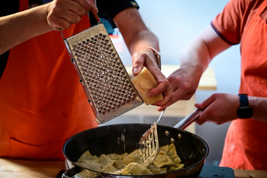 Zwei Personen in orangen Schürzen kochen zusammen: eine reibt Parmesan über Ravioli in der Pfanne, die andere rührt. Zu sehen sind nur Hände und Oberkörper.