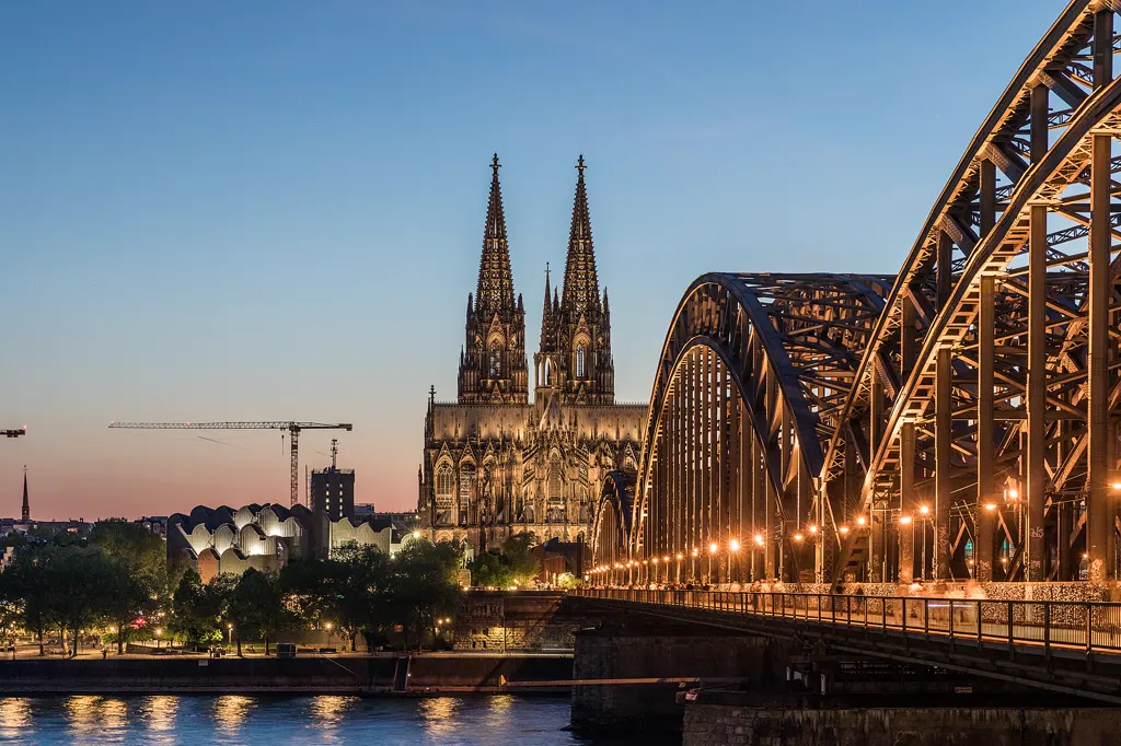 Kölner Dom mit der 2025 eingeweihten LED-Beleuchtung in der blauen Stunde, vom Deutzer Rheinufer aus gesehen; rechts die beleuchtete Hohenzollernbrücke, unten der Rhein, links am Horizont ein Baukran.
