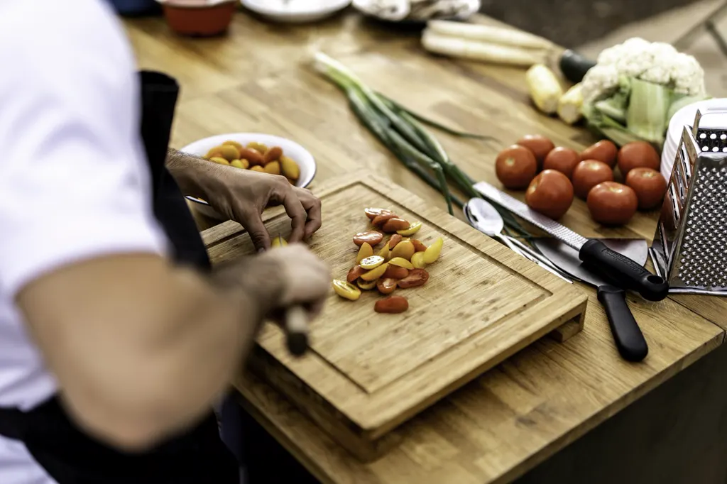 Hände schneiden Tomaten auf einem Holzbrett, daneben Lauchzwiebeln, Blumenkohl und Reibe