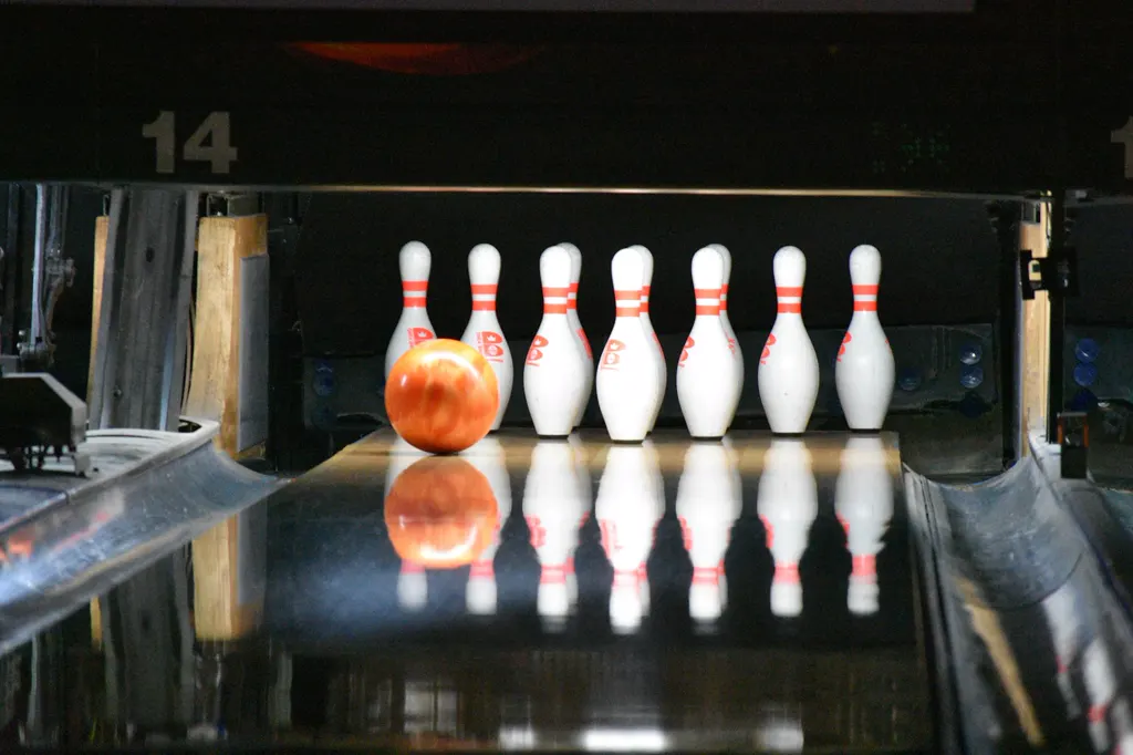 Eine orangefarbene Bowlingkugel rollt auf der glänzenden Bahn auf die weißen Pins zu, die sich im Holz spiegeln.