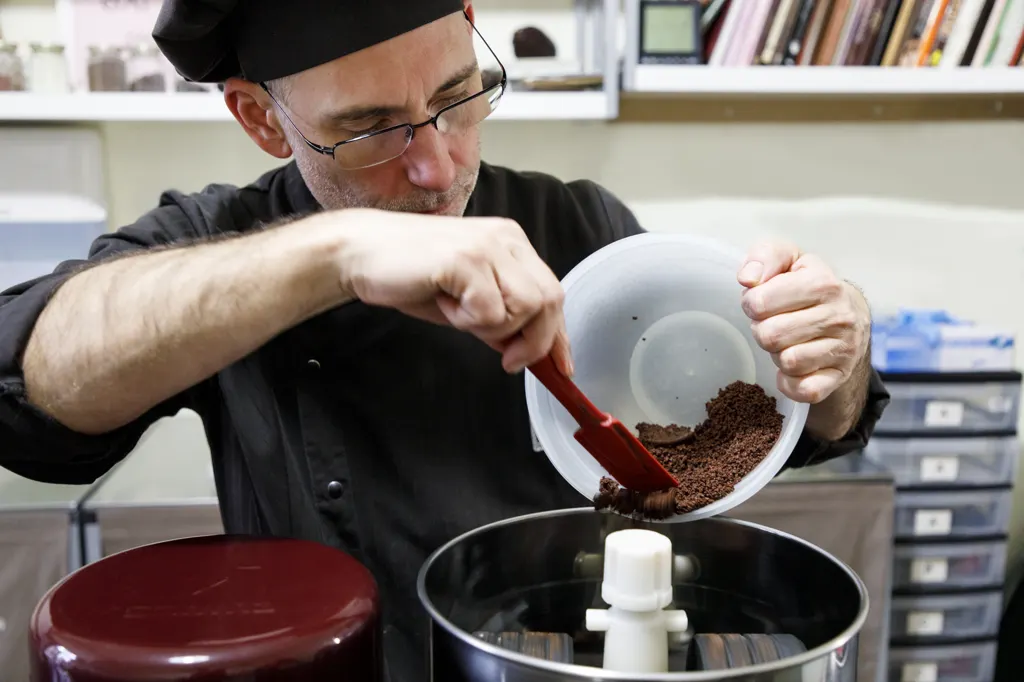 Chocolatier Michael Kitz füllt in der Backstube mit einem roten Spatel gemahlene Kakaobohnen in eine Edelstahlmaschine.