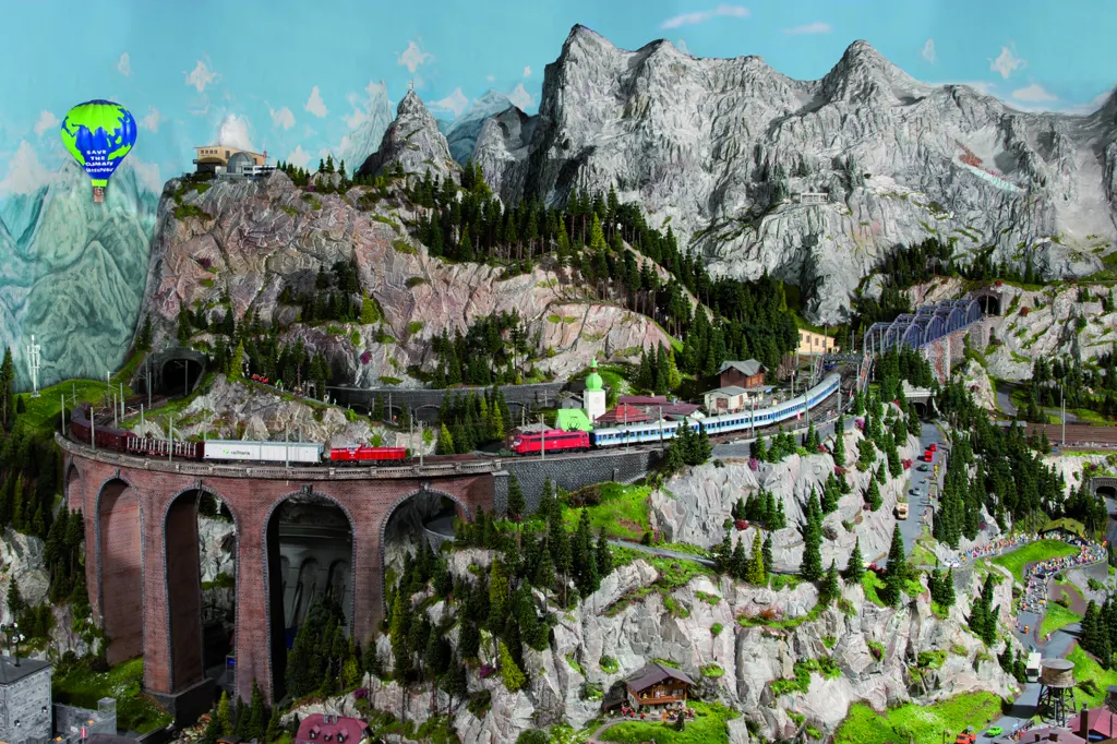 Modelleisenbahn im Österreich-Abschnitt: Züge auf einem Steinviadukt am Berghang, dahinter Felsgipfel und Nadelwald