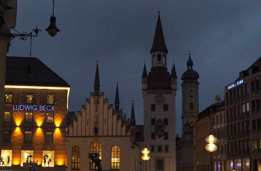 Altes Rathaus am Marienplatz mit Turm und Uhr am Abend, dahinter ein Kirchturm, alte Hängelaternen im Vordergrund; ohne Personen