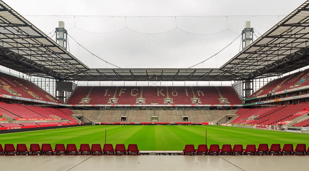 Leeres RheinEnergieSTADION vom Spielfeldrand aus: vorn eine Reihe roter Sitzschalen, dahinter der grüne Rasen und die gegenüberliegende Tribüne mit dem Sitzschriftzug „1. FC Köln“, darüber das Dach mit zwei Pylonen.