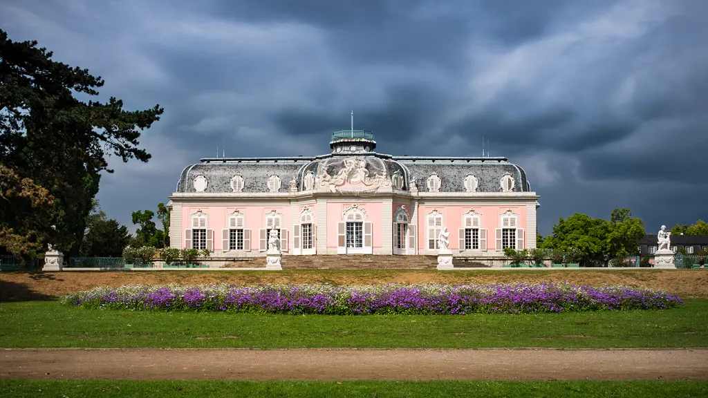 Corps de Logis von Schloss Benrath von der Gartenseite, sonnenbeschienen vor dunklem Wolkenhimmel, davor ein blühendes Beet