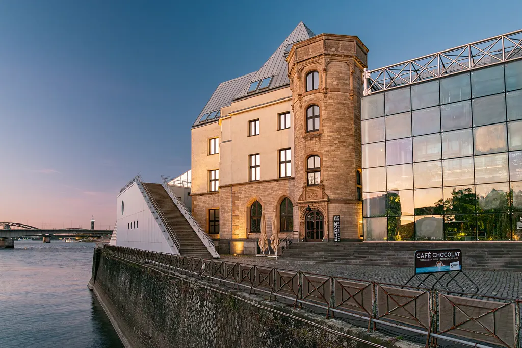 Das Schokoladenmuseum Köln am Rheinufer zur blauen Stunde, mit warm beleuchtetem Altbau samt Turm, weißem Museumsflügel mit Treppe und gläsernem Anbau.