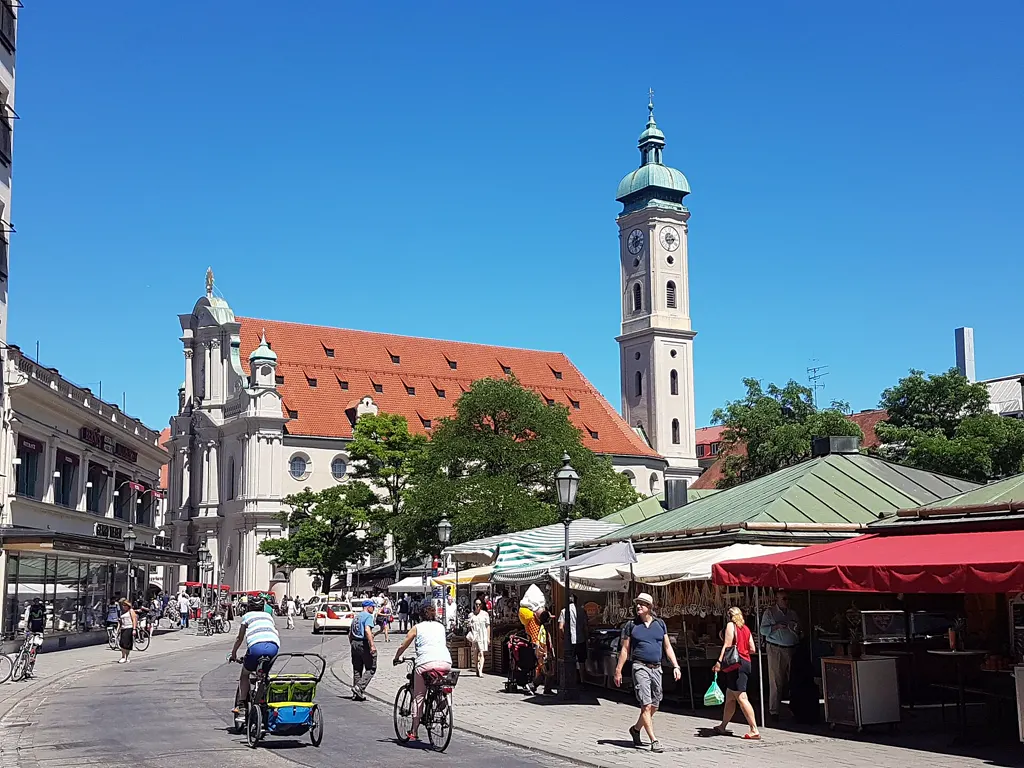Viktualienmarkt in München mit Marktständen unter grünen Dächern und Markisen, dahinter die Heilig-Geist-Kirche mit Turm bei blauem Himmel.