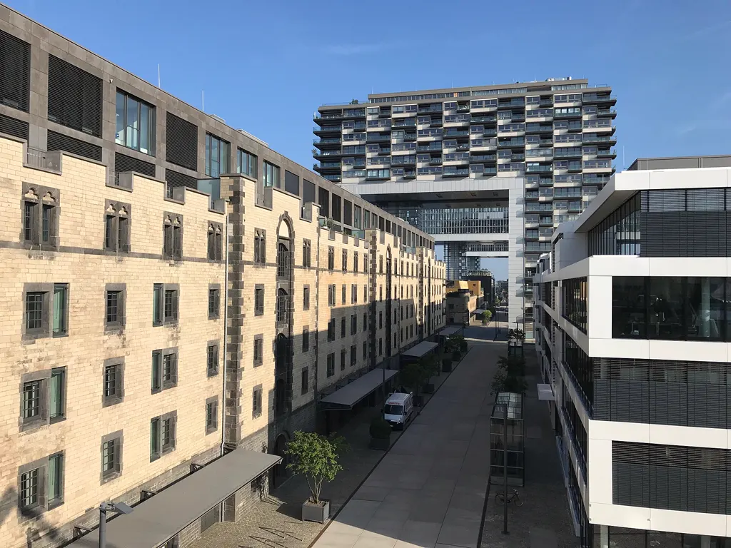 Straße im Kölner Rheinauhafen mit der langen historischen Halle 11 links, einem Kranhaus im Hintergrund und einem modernen Bürogebäude rechts.