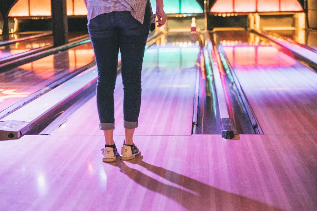 Person in Jeans und Bowlingschuhen, von hinten gesehen am Anlauf, davor glänzende Bahnen in Rosa-, Orange- und Türkistönen.