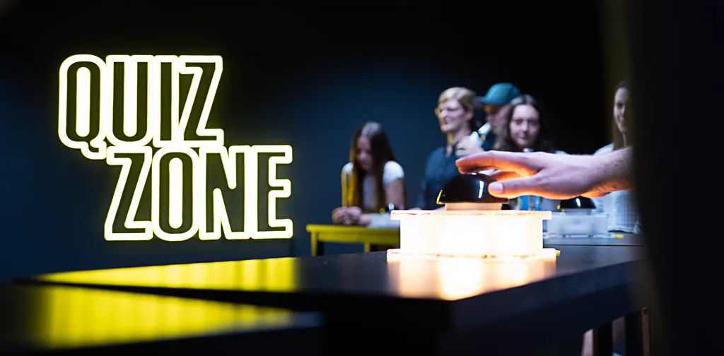 Hände am Buzzer vor der Leuchtschrift QUIZ ZONE